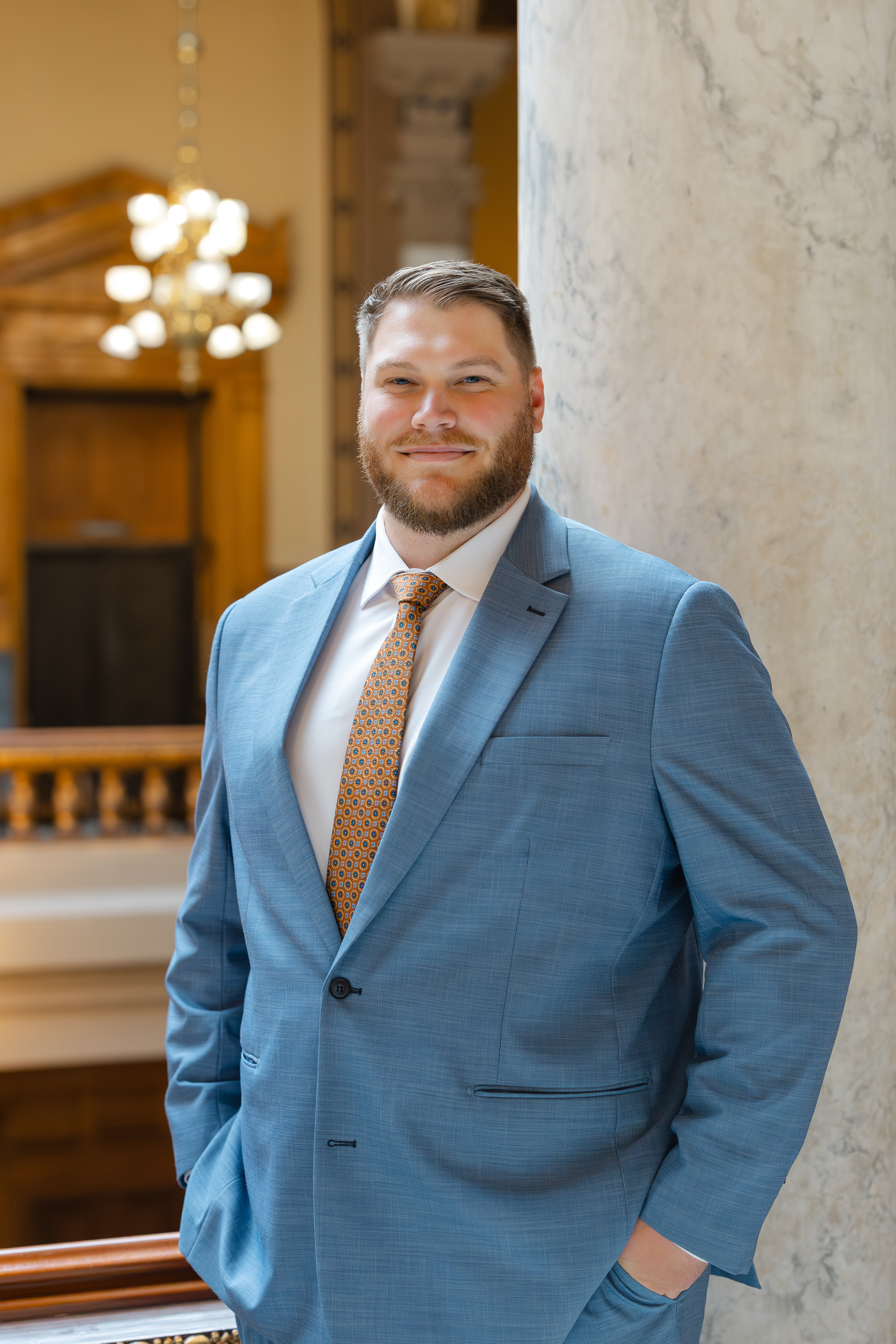 A well-dressed man with a beard poses in a light blue suit and patterned orange tie, standing against a marble pillar in a warmly lit interior.